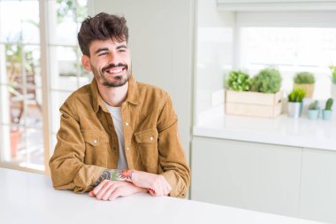 Beyaz masada oturan rahat ceket giyen genç adam yüzünde gülümseme ile yan bakmak, doğal ifade. Güven gülüyor.