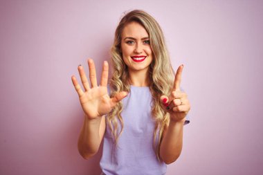 Pembe izole arka planda duran mor t-shirt ayakta giyen genç güzel kadın ve emin ve mutlu gülümseyen sırasında parmak numarası altı ile işaret.