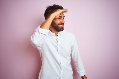Genç Hint işadamı izole pembe arka plan üzerinde ayakta zarif gömlek giyiyor çok mutlu ve gülümseyen çok uzağa baş el ile bakıyor. Arama konsepti.