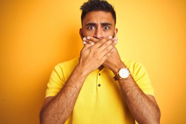 Izole sarı arka plan üzerinde duran polo giyen genç Hintli adam hata için elleri ile ağız kapsayan şok. Gizli kavram.