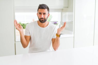 Yakışıklı İspanyol adam evde rahat beyaz t-shirt silah ve elleri kaldırdı ile clueless ve karışık ifade. Şüphe kavramı.