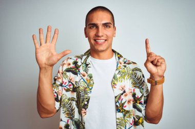 Beyaz arka planda gösteren ve parmak sayısı altı ile işaret yukarı üzerinde Hawaii gömlek giyen tatil genç yakışıklı adam emin ve mutlu gülümseyen iken.