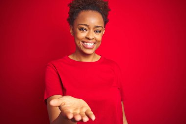 Young beautiful african american woman with afro hair over isolated red background smiling cheerful offering palm hand giving assistance and acceptance. clipart