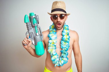 Genç adam yaz şapka giyen ve Hawaii Lei çiçek izole arka plan üzerinde şok bir sürpriz yüz ile korkuyor ve korku ifadesi ile heyecanlı korkmuş üzerinde su tabancası tutan
