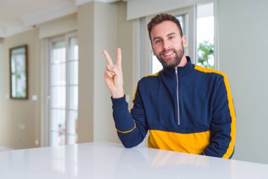 Yakışıklı adam evde mutlu yüz zafer işareti yaparken kameraya göz kırpıyor ile gülümseyen. İki numaralı.