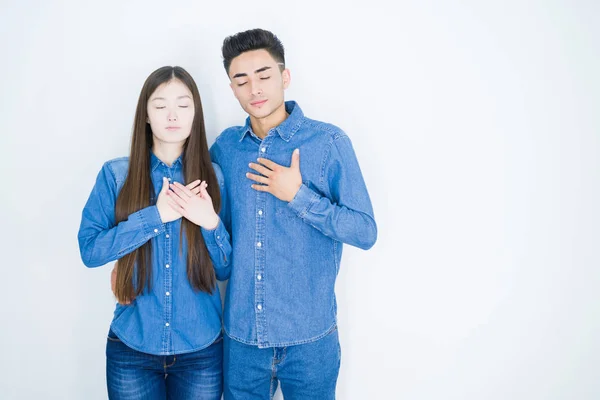 Beautiful young asian couple over white isolated background smiling with hands on chest with closed eyes and grateful gesture on face. Health concept.