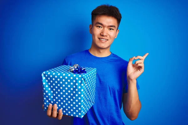 Young asian chinese man holding birthday gift standing over isolated blue background very happy pointing with hand and finger to the side
