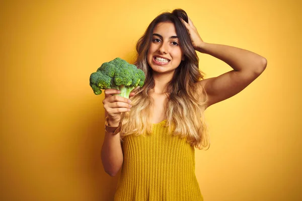 黄色の孤立した背景の上にブロッコリーを食べる若い美しい女性は 頭の上に手で強調し 恥と驚きの顔でショックを受け 怒りとイライラ 間違いに対する恐怖と動揺 — ストック写真