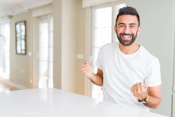 ハンサムなヒスパニック系の男性カジュアルな白い シャツ自宅非常に幸せと興奮の両手を上げると 勝者のジェスチャーをして笑みを浮かべて 成功のために叫んでいます お祝いのコンセプト — ストック写真