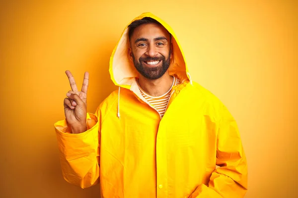 孤立した黄色の背景の上にフード付きのレインコートを着た若いインド人男性は 勝利のサインをしているカメラで幸せそうな顔をして微笑んでいます ナンバー2 — ストック写真