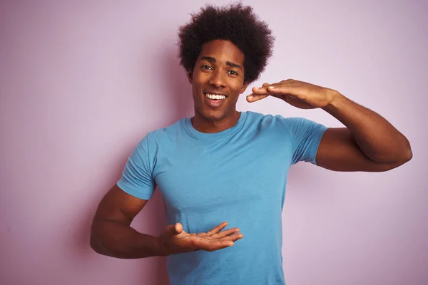 African american man with afro hair wearing blue t-shirt standing over isolated pink background gesturing with hands showing big and large size sign, measure symbol. Smiling looking at the camera. Measuring concept.