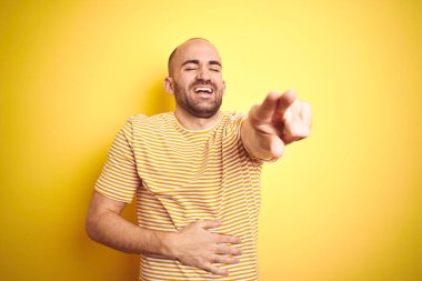 Sarı izole arka planda size gülüyor üzerinde rahat çizgili t-shirt giyen sakal ile genç kel adam, vücut üzerinde el ile kameraya parmak işaret, utanç ifadesi