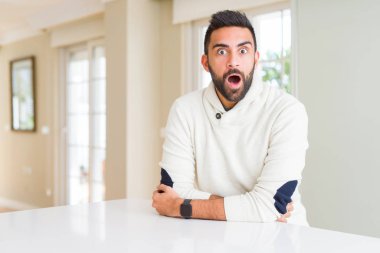 Evde rahat beyaz kazak giyen yakışıklı İspanyol adam korkuyor ve sürpriz ifade, korku ve heyecanlı yüz ile şok.