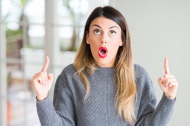 Evde kış kazak giyen genç güzel kadın hayrete ve yukarıya bakıyor ve parmakları ile işaret ve silah kaldırdı şaşırttı.