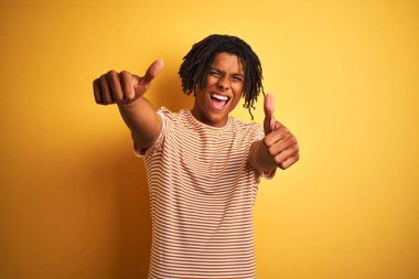 Dreadlocks ile Afro adam izole sarı arka plan üzerinde duran çizgili t-shirt ayakta, el ile olumlu jest yapıyor onaylar, gülümseyen ve başarı için mutlu kadar başparmak. Kazanan jest.