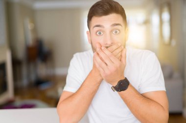 Evde rahat beyaz t-shirt giyen genç yakışıklı adam yanlışlık için eller ile ağız kapsayan şok. Gizli konsept.