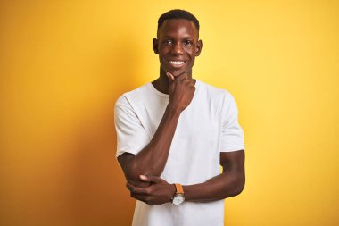 Beyaz tişört giyen genç Afrikalı Amerikalı adam izole sarı arka plan üzerinde ayakta silah ve el çene üzerinde yükseltilmiş ile gülümseyen kamera emin bakıyor. Pozitif düşünme.