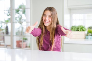 Beyaz masada güzel genç kız çocuk el ile olumlu jest yapıyor onayıyla, gülümseyen ve başarı için mutlu kadar başparmak. Kamera bakarak, kazanan jest.
