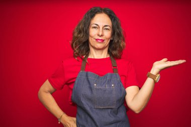 Orta yaş kıdemli kadın kırmızı izole arka plan gülümseyen neşeli sunumu üzerinde apron üniforma giyiyor ve kamera bakarak el avuç ile işaret.