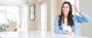 Evde beyaz masada oturan güzel genç kadının geniş açı resmi el ve parmaklar ile ok işareti yapıyor pozitif gülümseyen. Başarılı ifade.