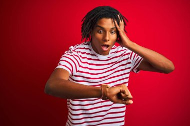 Genç Afro adam ile dreadlocks giyen çizgili t-shirt ayakta üzerinde izole kırmızı arka plan saat zaman endişeli, geç elde korkuyor bakıyor