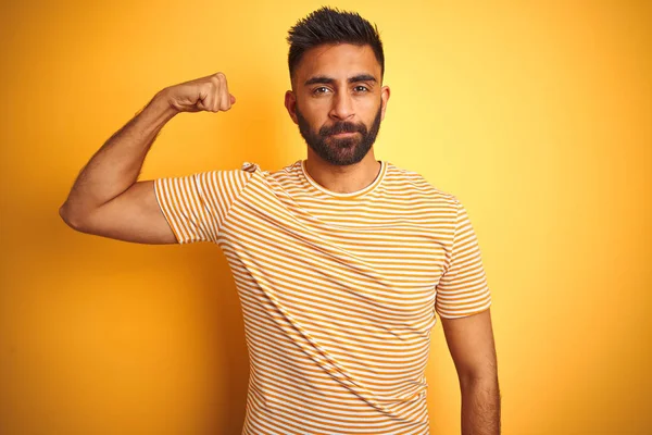 Young indian man wearing t-shirt standing over isolated yellow background Strong person showing arm muscle, confident and proud of power