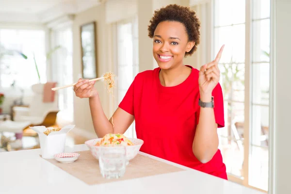 Young african american woman with afro hair eating asian food at home very happy pointing with hand and finger to the side