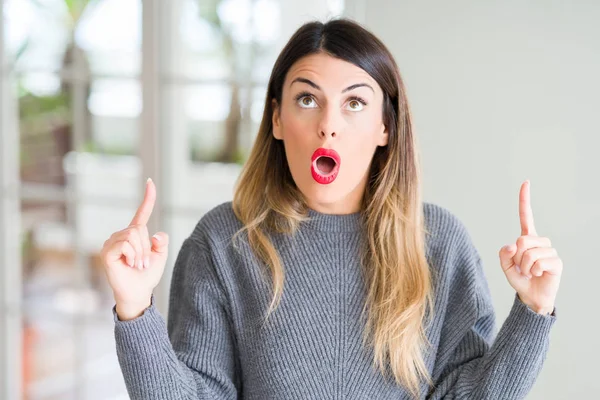 Evde kış kazak giyen genç güzel kadın hayrete ve yukarıya bakıyor ve parmakları ile işaret ve silah kaldırdı şaşırttı.