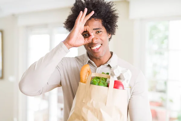 African American man holding groceries bag with fresh vegetables at home with happy face smiling doing ok sign with hand on eye looking through fingers