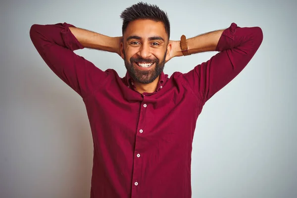 Young indian man wearing red elegant shirt standing over isolated grey background relaxing and stretching, arms and hands behind head and neck smiling happy