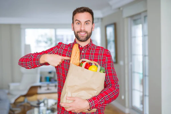 Handsome man holding groceries bag with surprise face pointing finger to himself