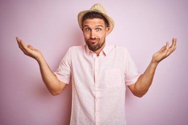 Yakışıklı adam pembe izole arka plan üzerinde şapka ve yazlık gömlek giyen silah ve elleri kaldırdı ile anlamsız ve karışık ifade. Şüphe kavramı.