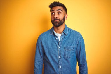 İzole sarı arka plan üzerinde ayakta kot gömlek giyen genç Hintli adam yan bakıyor ve düşünmek uzağa bakıyor.