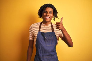 Afro Amerikan barista adam ile dreadlocks giyen apron üzerinde izole sarı arka plan yapıyor mutlu thumbs yukarı jest ile el. Başarı göstererek kameraya bakarak ifadeyi onaylama.