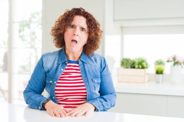 Evde kot ceket giyen kıvırcık saçlı orta yaş yaşlı kadın Şok yüzünde, şüpheci ve alaycı görünümlü, açık ağız ile şaşırttı
