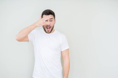 Mahcup ifadeyle parmaklarımın arasından seyir yüz ve gözler el ile kapsayan şok bakmak izole arka plan üzerinde rahat beyaz t-shirt giyen genç yakışıklı adam.