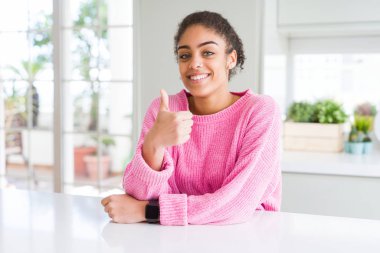 Afro saç ile güzel Afrikalı Amerikan kadın rahat pembe kazak el ile jest kadar mutlu thumbs yapıyor giyiyor. Başarı gösteren kameraya bakarak ifadeyi onaylama.