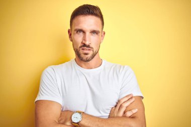 Sarı izole arka plan üzerinde rahat beyaz t-shirt giyen genç yakışıklı adam şüpheci ve sinir, Çapraz kollar ile yüz üzerinde ifade onaylamıyor. Negatif kişi.