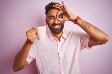 Genç Hint adam giymek gömlek içme fincan arasında kahve Standing üzerinde izole pembe arka plan ile mutlu yüz gülümseyen yapıyor ok işareti ile göz üzerinde görünümlü parmak