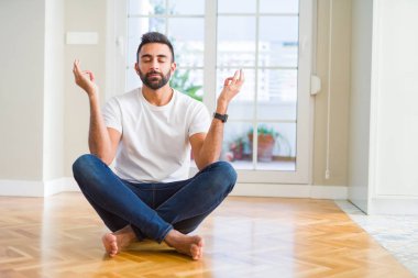 Rahat t-shirt evde yerde oturan giyen yakışıklı İspanyol adam Relax ve gözler ile gülümseyen parmaklar ile meditasyon jest yapıyor kapalı. Yoga konsepti.
