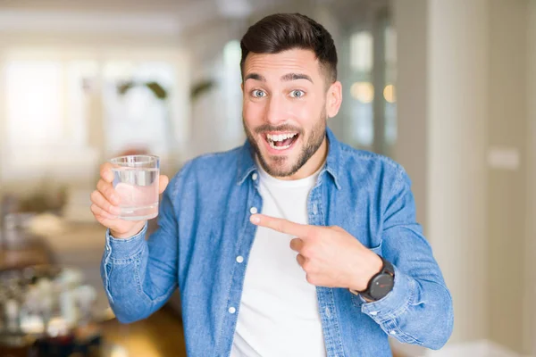 自宅で水のコップを飲む若いハンサムな男は 手と指で指して非常に幸せ — ストック写真