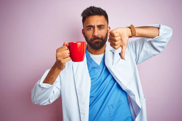 Young indian doctor man drinking cup of coffee standing over isolated pink background with angry face, negative sign showing dislike with thumbs down, rejection concept