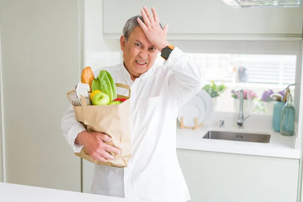 Handsome senior man holding a paper bag of fresh groceries at the kitchen stressed with hand on head, shocked with shame and surprise face, angry and frustrated. Fear and upset for mistake.