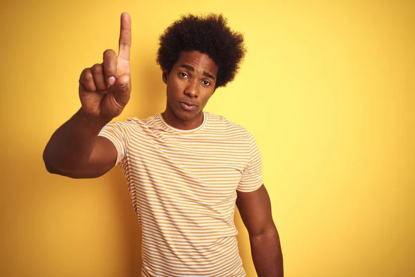 American man with afro hair wearing striped t-shirt standing over isolated yellow background Pointing with finger up and angry expression, showing no gesture