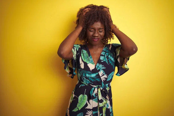若いアフリカのアフロ女性は 痛みと偏頭痛のために絶望的な頭痛に苦しんで孤立した黄色の背景の上に夏の花のドレスを着て 頭の上に手を置く — ストック写真
