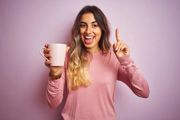 年轻的漂亮女人拿着一杯咖啡在粉红色孤立的背景惊讶与一个想法或问题指向手指与快乐的脸 — 图库照片