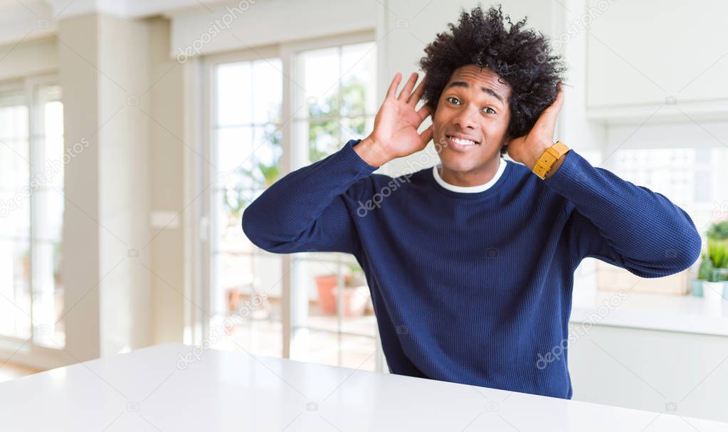 Young african american man wearing casual sweater sitting at home Trying to hear both hands on ear gesture, curious for gossip. Hearing problem, deaf