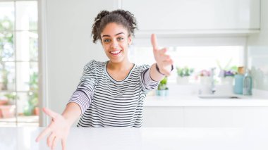 Afro saç ile güzel Afrikalı Amerikan kadın rahat çizgili kazak kucaklamak için açık kollar ile gülümseyen kamera bakıyor giyiyor. Mutluluğu kucaklayan neşeli ifade.