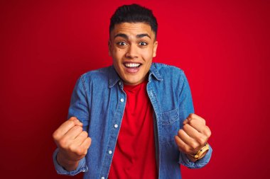İzole kırmızı arka plan üzerinde ayakta kot gömlek giyen genç Brezilyalı adam şaşırttı kutluyor ve başarı için hayran silah kaldırdı ve açık gözleri ile. Kazanan kavramı.