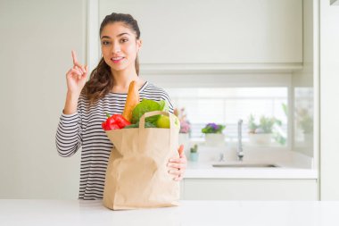 Yiyecek dolu kağıt torbası tutan genç kadın bir fikir veya soru mutlu yüz, bir numara ile parmak işaret ile şaşırdı